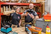 Een man en een vrouw (links) kijken lachend in de camera. De vrouw draagt een lange dunne balk op haar schouder en de man heeft een lijmpistool vast. Ze staan achter een werkbank, en achter hun staat een groot metalen rek met diverse soorten planken.